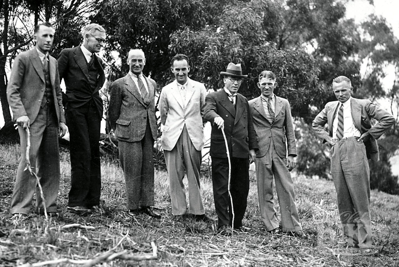 Black-and-white photograph taken at Bathurst in 1937, showing six men in suits standing on a grassy hillside. Motorcycle champions Stanley Woods and Don Bain stand on either side of Mayor Martin Griffin's walking stick, with trees and bushland in the background.